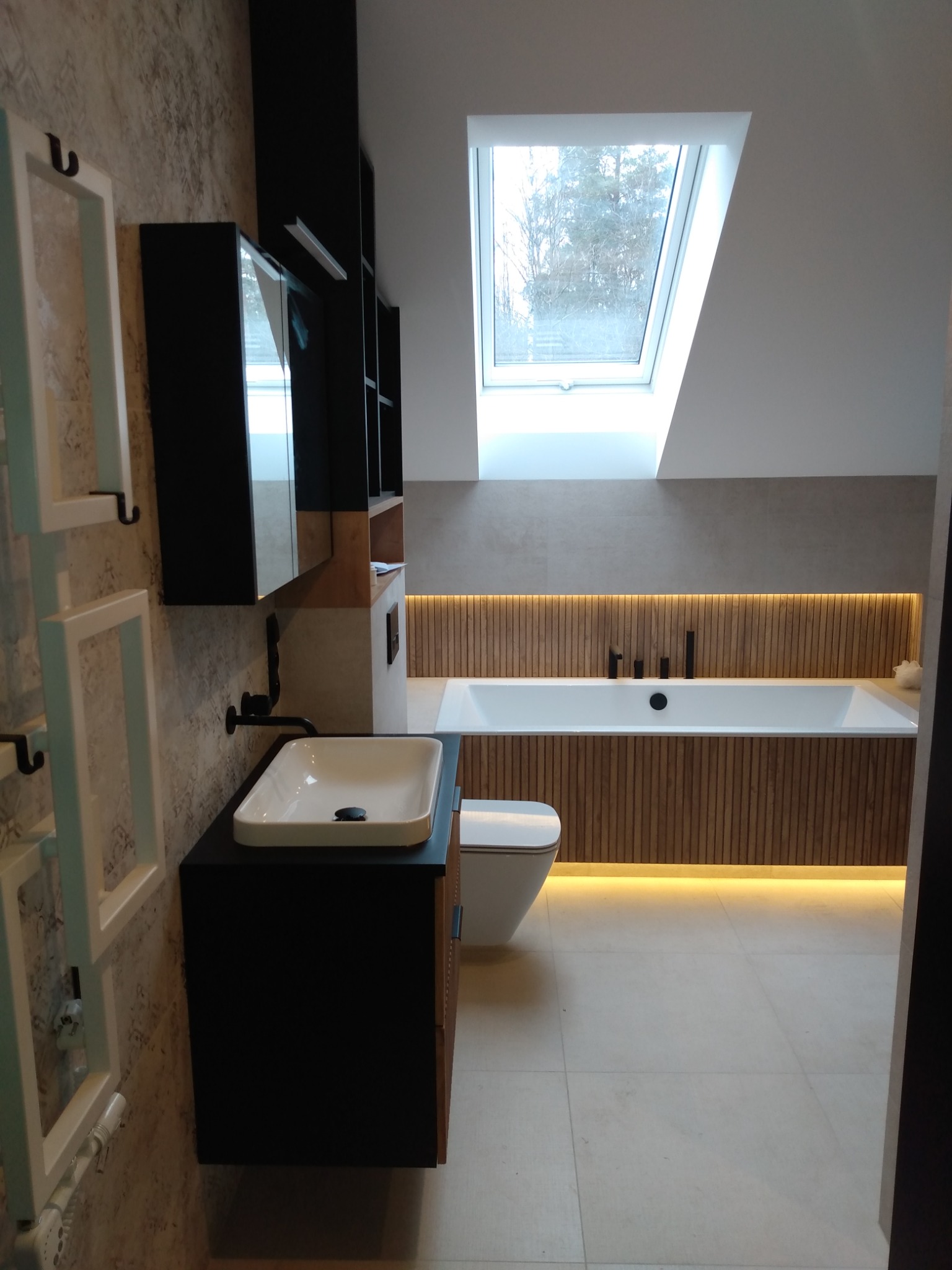 Nowoczesna łazienka na poddaszu z oknem dachowym, umywalką nablatową na czarnej szafce, podświetlaną wanną obudowaną drewnianymi panelami oraz grzejnikiem drabinkowym.