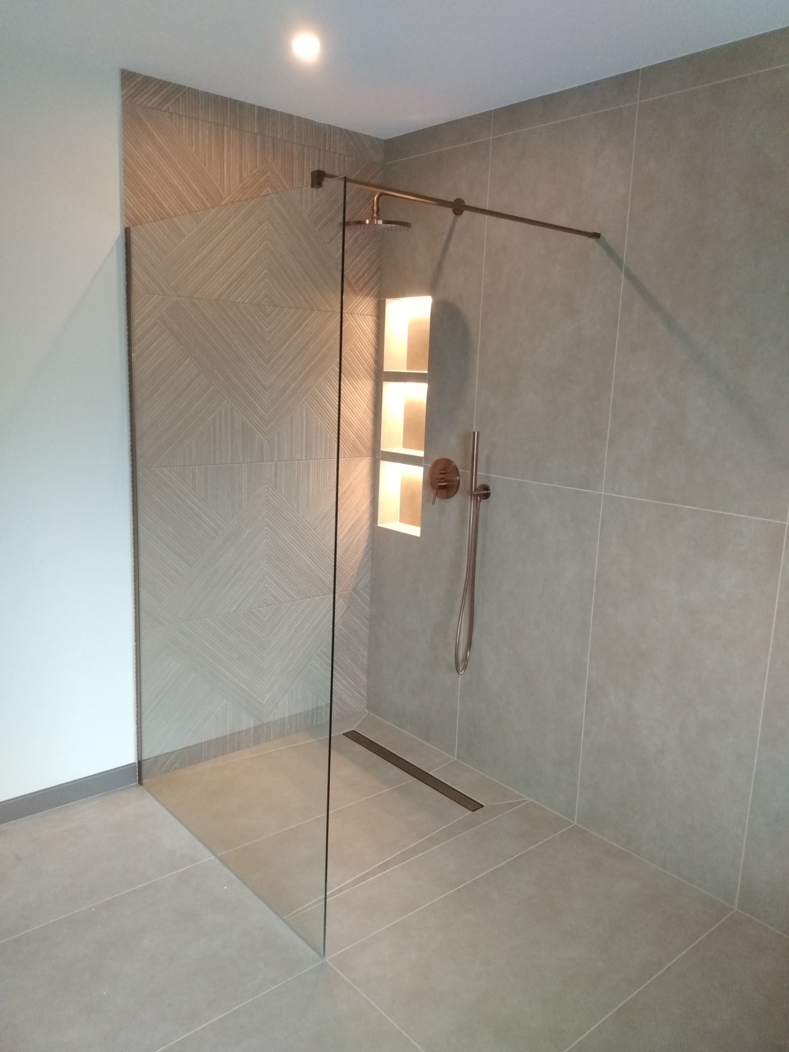 Nowoczesna kabina prysznicowa walk-in z szklaną ścianką, wykończona płytkami w odcieniach szarości, z dekoracyjną ścianką z geometrycznym wzorem i wnęką oświetloną od wewnątrz.