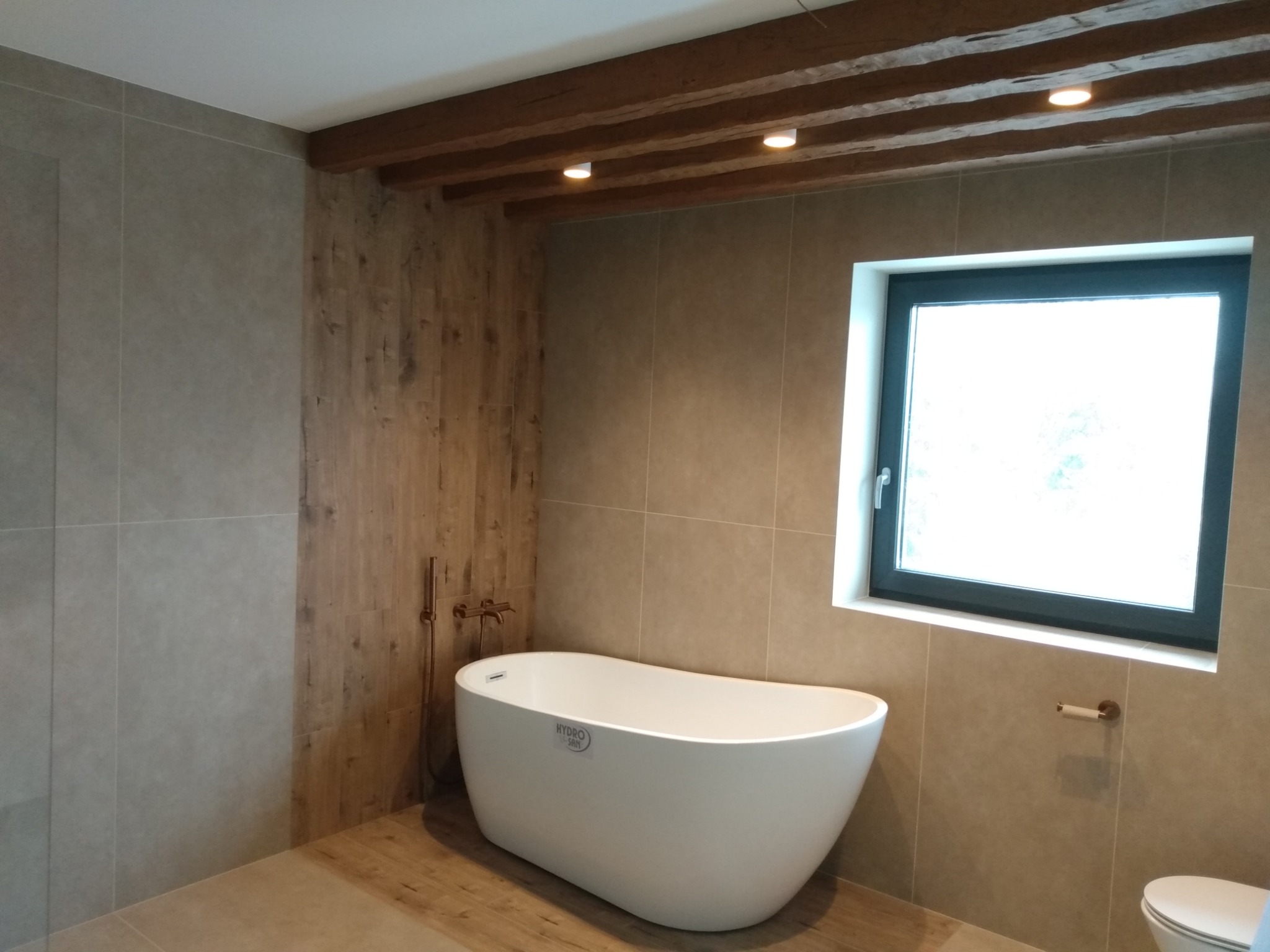 Nowoczesna łazienka z wolnostojącą białą wanną, drewnianą ścianą i sufitem z belkami, oświetlona punktowymi lampami, z oknem w czarnej ramie.