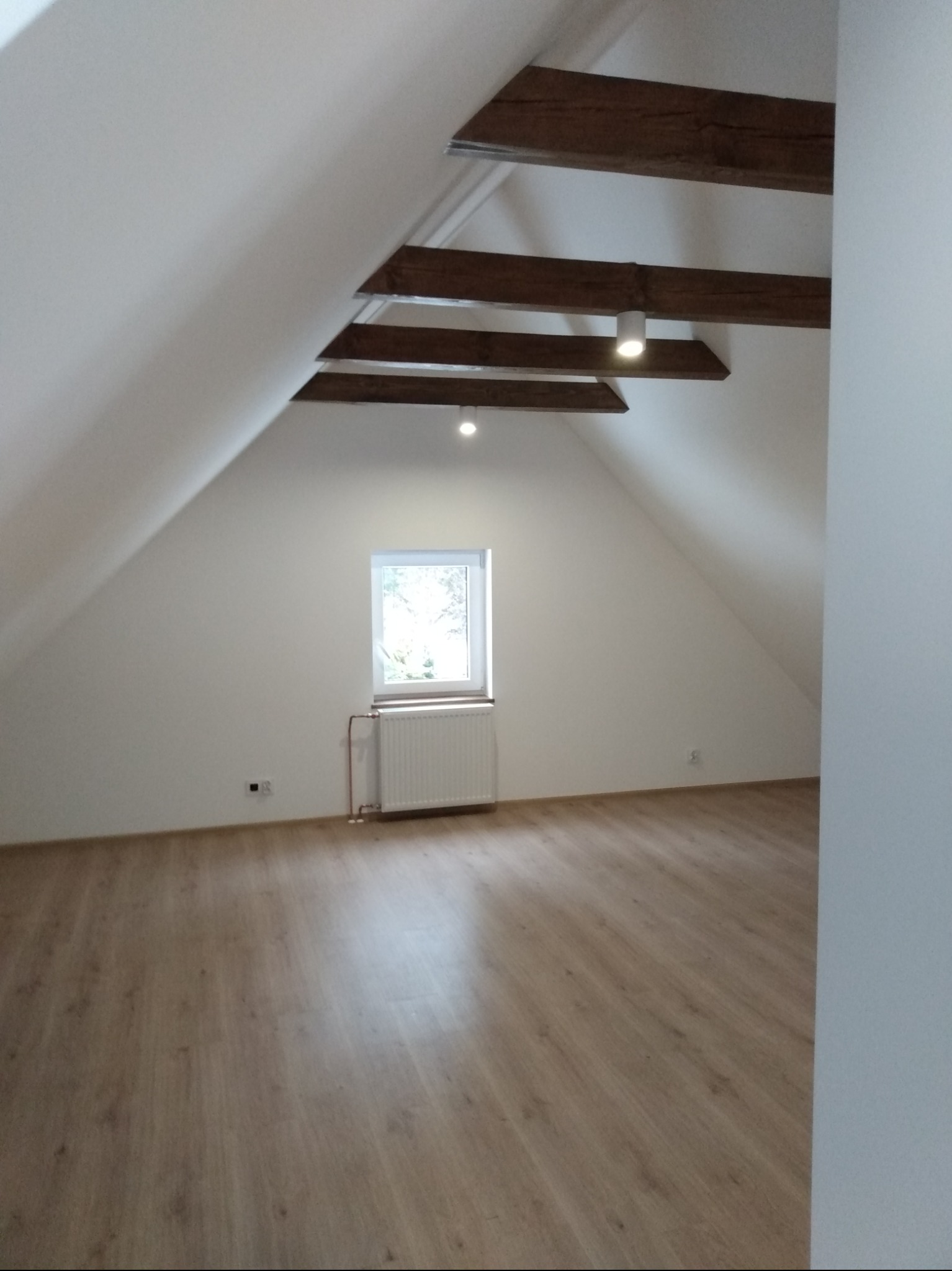 Poddasze po remoncie z drewnianymi belkami, oknem i panelami podłogowymi w odcieniach brązu.