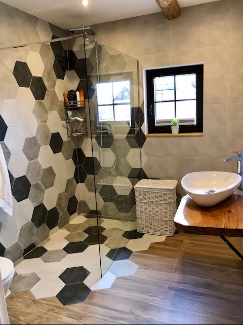 Nowoczesna łazienka z kabiną prysznicową i heksagonalnymi płytkami w odcieniach szarości i bieli, drewnianą umywalką i wiklinowym koszem na pranie.