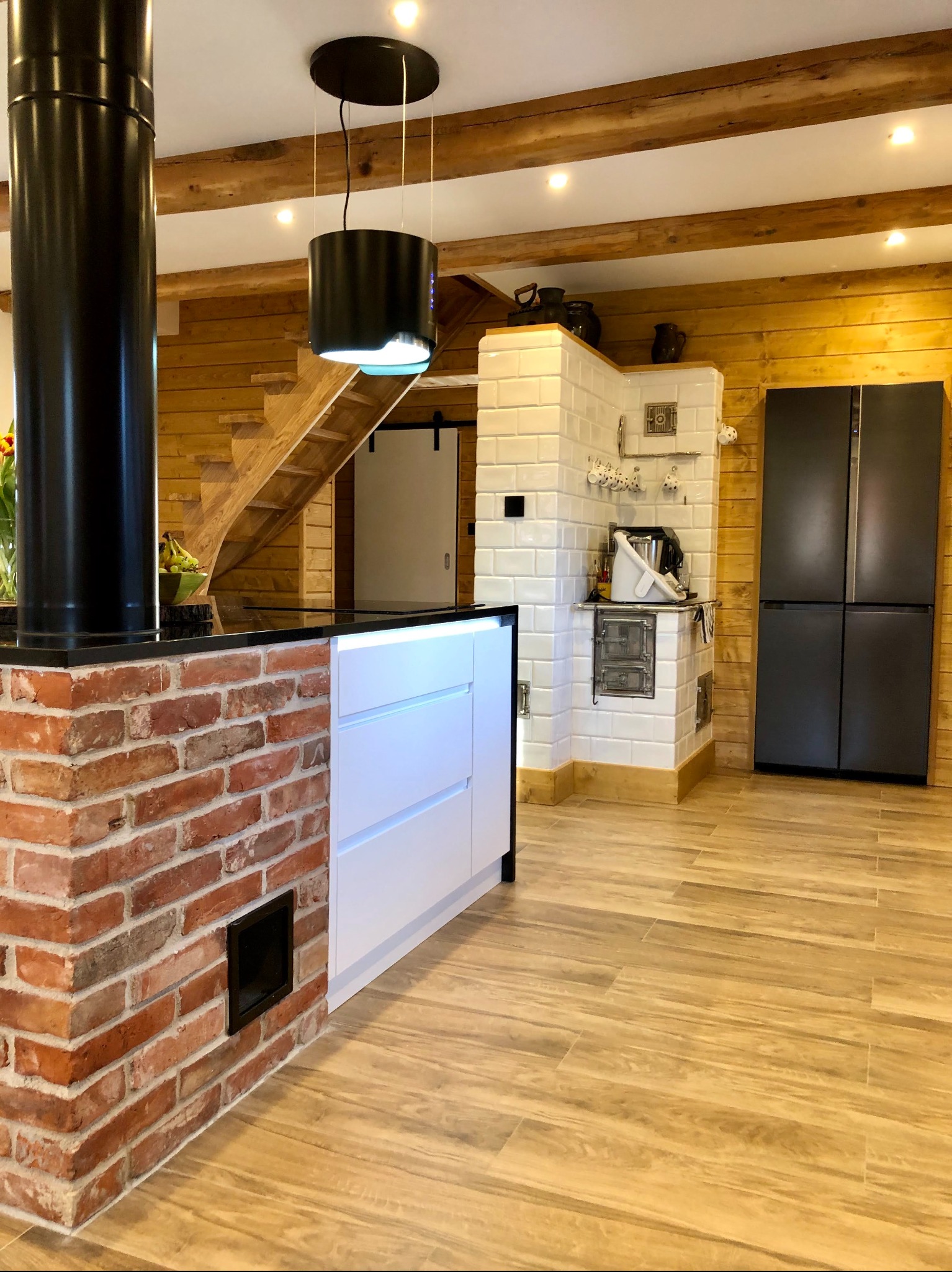 Kuchnia w stylu rustykalnym z drewnianymi ścianami, ceglaną zabudową kominkową z czarnym kominem, białą zabudową meblową, piecem kaflowym i czarną lodówką.