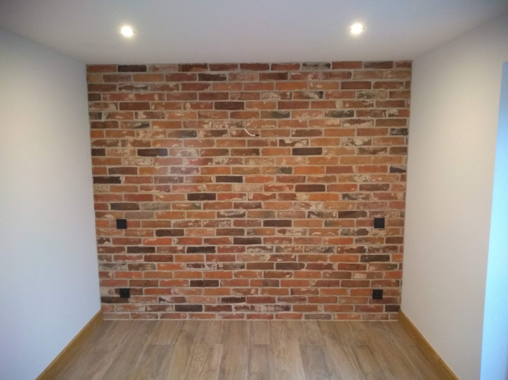 Ściana wykończona cegłą dekoracyjną w odcieniach brązu i czerwieni, z widocznymi puszkami elektrycznymi, podświetlona dwoma punktowymi lampami sufitowymi, z drewnianą podłogą w jasnym odcieniu.