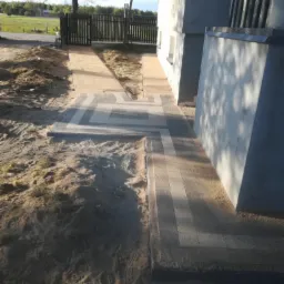 Świeżo ułożona, dwukolorowa kostka brukowa prowadząca od furtki w drewnianym ogrodzeniu do ściany budynku, otoczona piaskiem.