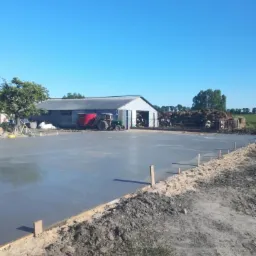 Świeżo wylana, gładka posadzka betonowa na zewnątrz budynku gospodarczego z widocznym traktorem i krowami w tle, w słoneczny dzień.