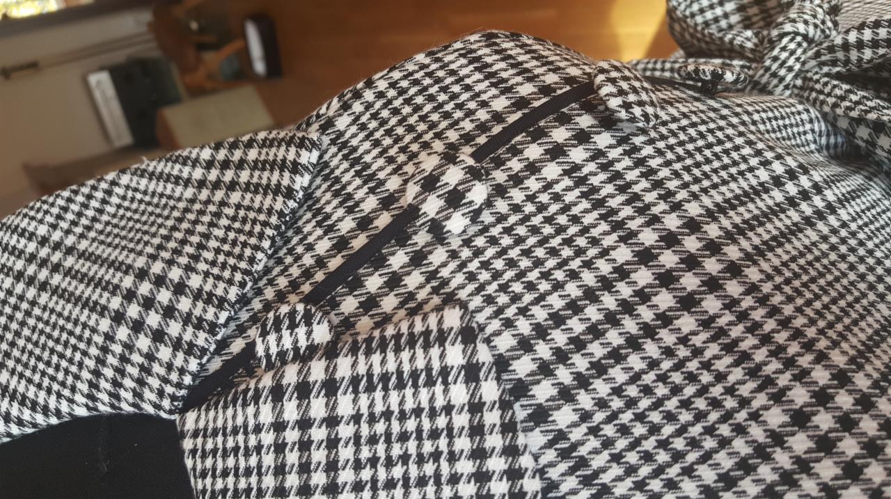 Fragment marynarki w pepitkę z czarną lamówką przy klapie, widoczny fragment podszewki w dolnej części kadru.