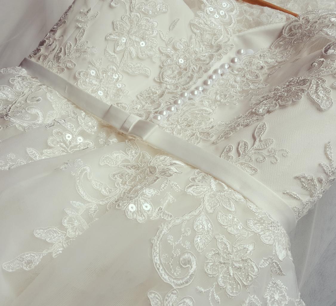 Fragment sukni ślubnej z koronkowymi aplikacjami, perełkami i satynową szarfą z kokardką, widoczne detale zdobień.