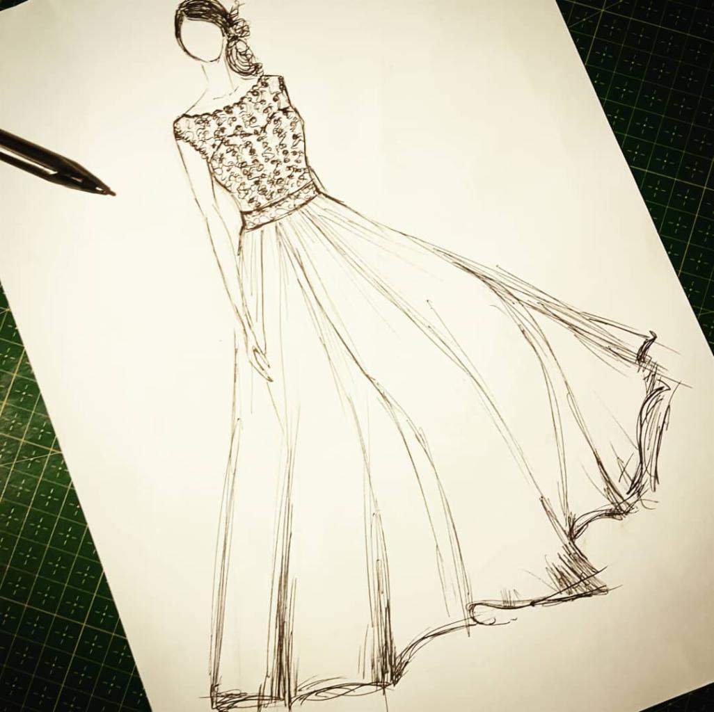Szkic sukni wieczorowej z koronkową górą i zwiewnym dołem, narysowany czarnym cienkopisem na białym papierze, leżącym na zielonej macie do cięcia.