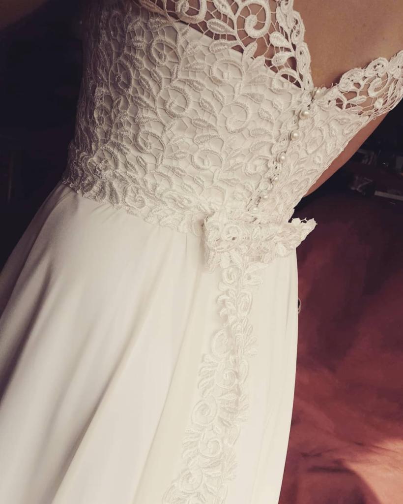 Delikatna, biała suknia z koronkową górą i zdobieniami z perełek, widoczna z bliska, z nieostrym tłem w odcieniach bordo.