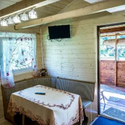 Wnętrze drewnianego domku letniskowego: stół nakryty obrusem z wzorem, narożna kanapa ze wzorem w paski, telewizor na ścianie z desek, okno z firanką i widok na zewnątrz przez otwarte drzwi.