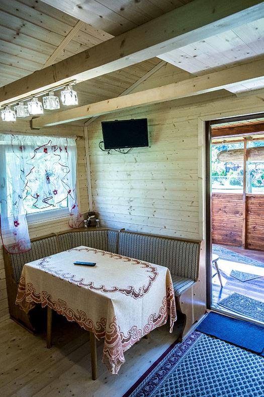 Wnętrze drewnianego domku letniskowego: stół nakryty obrusem z wzorem, narożna kanapa ze wzorem w paski, telewizor na ścianie z desek, okno z firanką i widok na zewnątrz przez otwarte drzwi.