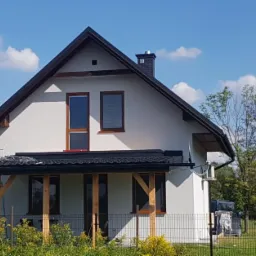 Biały dom jednorodzinny z ciemnym dachem i drewnianymi detalami okiennymi, widoczna drabina oparta o ścianę, błękitne niebo z chmurami w tle.