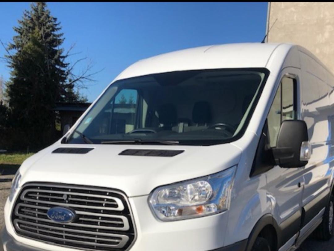 Biały samochód dostawczy Ford Transit z widocznym grillem i logo Forda, zaparkowany na zewnątrz w słoneczny dzień.
