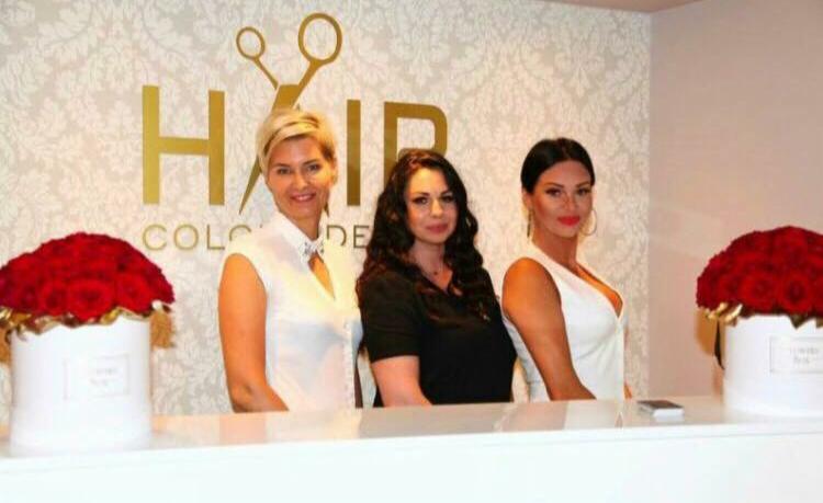 Trzy kobiety w recepcji salonu z logo z nożyczkami, dekoracjami z czerwonych róż w białych pudełkach i wzorzystą tapetą w tle.