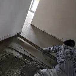 Pracownik w kombinezonie wyrównuje mokrą wylewkę jastrychową drewnianą łatą w jasnym pomieszczeniu z oknem.