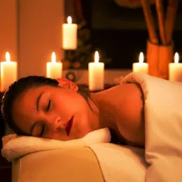 Kobieta leży na stole do zabiegów, przykryta białym prześcieradłem, z zamkniętymi oczami i czerwonymi ustami, otoczona blaskiem świec.