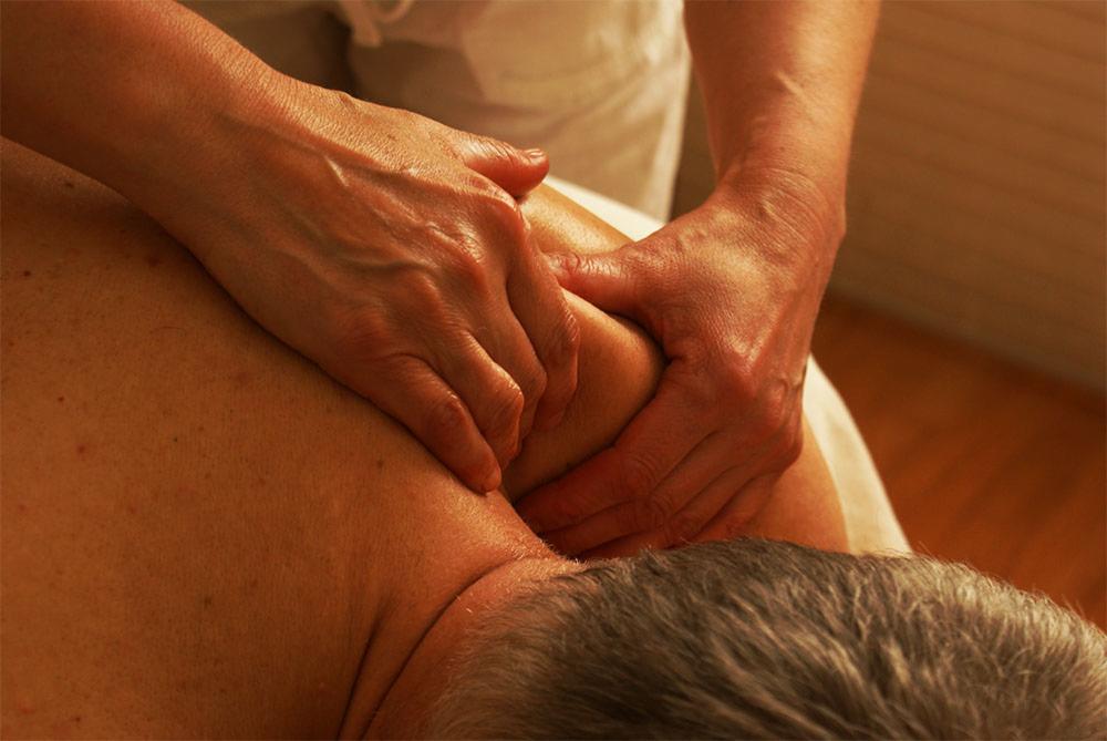 Masażysta ugniata bark pacjenta dwiema rękami, widoczna skóra i siwe włosy pacjenta, białe prześcieradło i drewniana podłoga w tle.