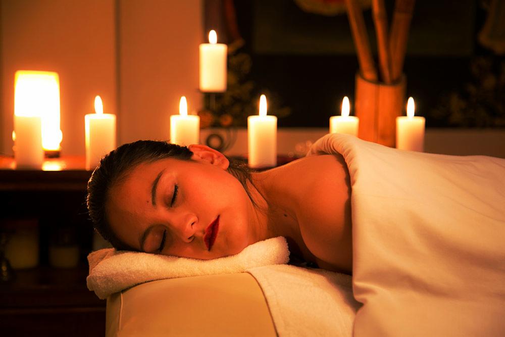 Kobieta leży na stole do zabiegów, przykryta białym prześcieradłem, z zamkniętymi oczami i czerwonymi ustami, otoczona blaskiem świec.