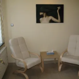 Jasny gabinet z dwoma fotelami, stolikiem z chusteczkami i obrazem kobiety z zasłoniętą twarzą na ścianie.