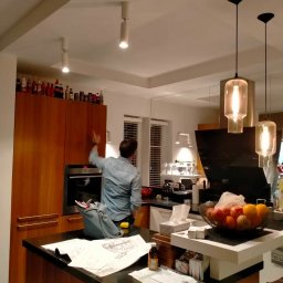 Project Group - Marzenna Cichocka - Mężczyzna w kuchni, prawdopodobnie podczas remontu, sprawdza górne szafki. Na blacie leżą plany, a w koszu owoce. Widoczne nowoczesne lampy.