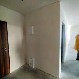 Project Group - Marzenna Cichocka - Wnętrze mieszkania w stanie deweloperskim: surowe ściany, betonowa podłoga, widoczne instalacje. Drzwi z ciemnego drewna i fragment osoby w żółtej kurtce w tle.