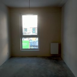 Project Group - Marzenna Cichocka - Surowe wnętrze pokoju z oknem i grzejnikiem. Widoczne ślady po pracach wykończeniowych na podłodze. Kabel zwisa z sufitu. Okno z widokiem na budowę.
