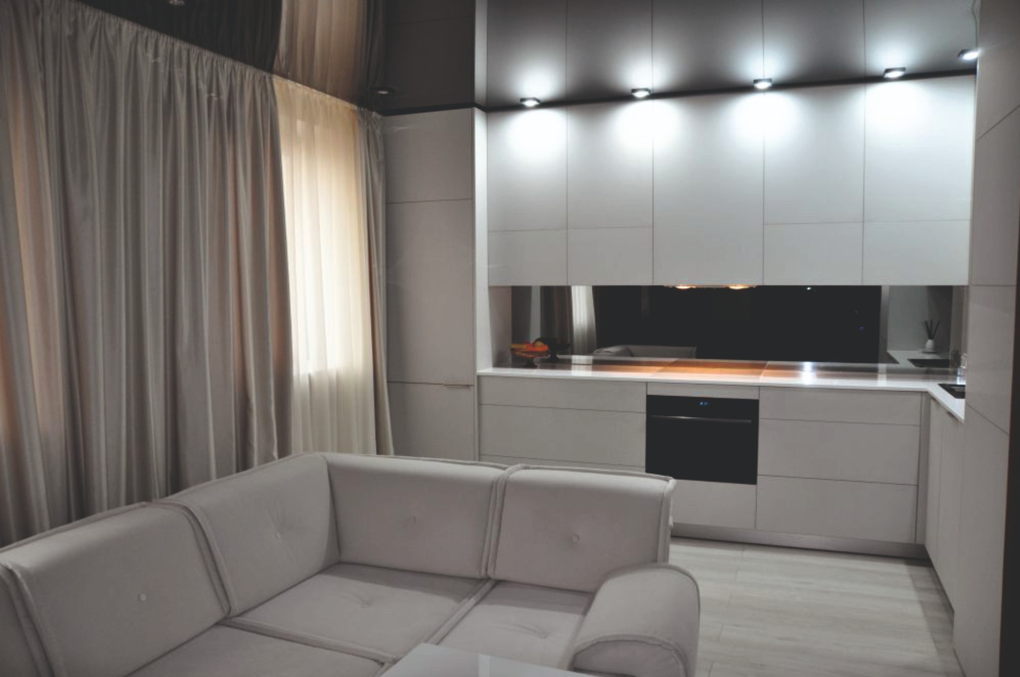 Minimalistyczna aranżacja wnętrza: szare zasłony, sofa i zabudowa kuchenna z oświetleniem punktowym. Nowoczesny design, jasne kolory, gładkie powierzchnie.