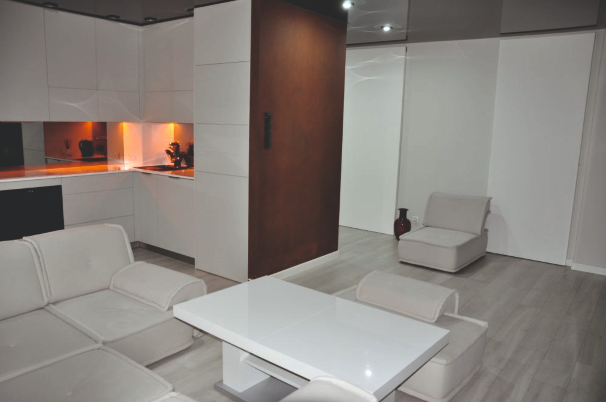 Minimalistyczne wnętrze: biała kuchnia z pomarańczowym akcentem, brązowa ściana, jasna sofa i stół. Nowoczesny design z drewnianą podłogą i dekoracyjnym wazonem.