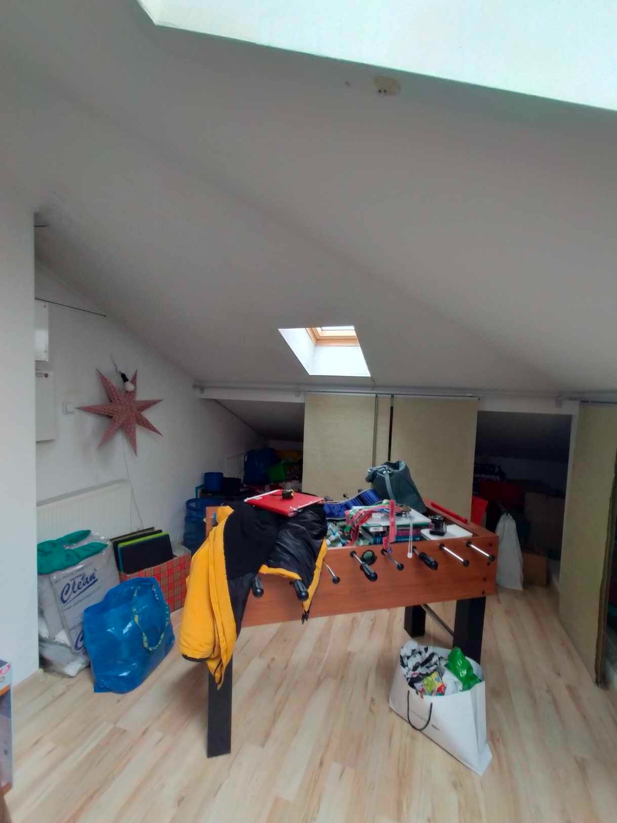 Poddasze użytkowe z oknem dachowym i stołem do piłkarzyków. Widoczne skosy, panele podłogowe i dekoracja w kształcie gwiazdy. Wnętrze po remoncie.