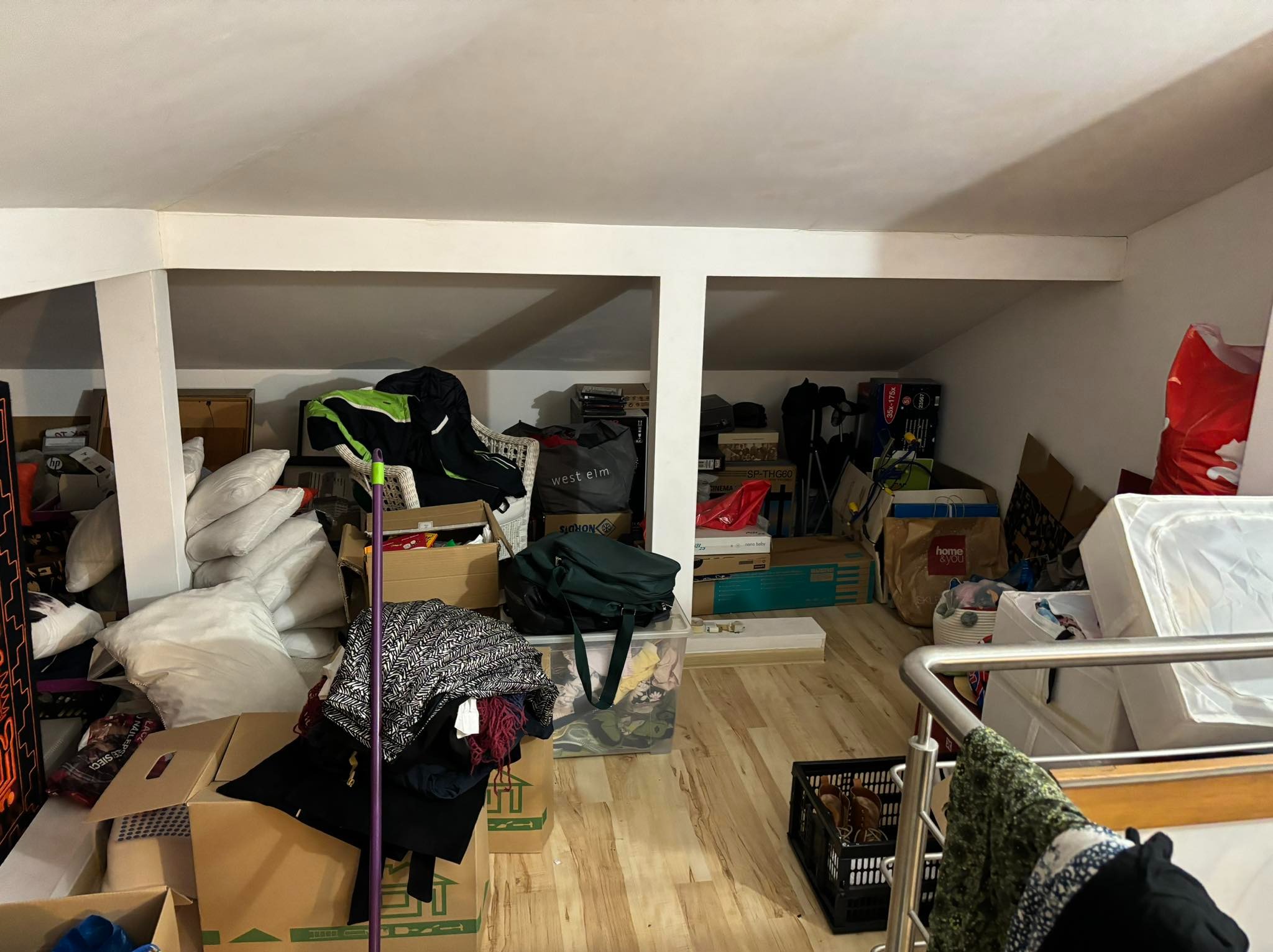 Poddasze użytkowe przed remontem: zagracona przestrzeń z kartonami, workami i meblami. Widoczne skosy dachu i drewniana podłoga. Chaos przed metamorfozą.