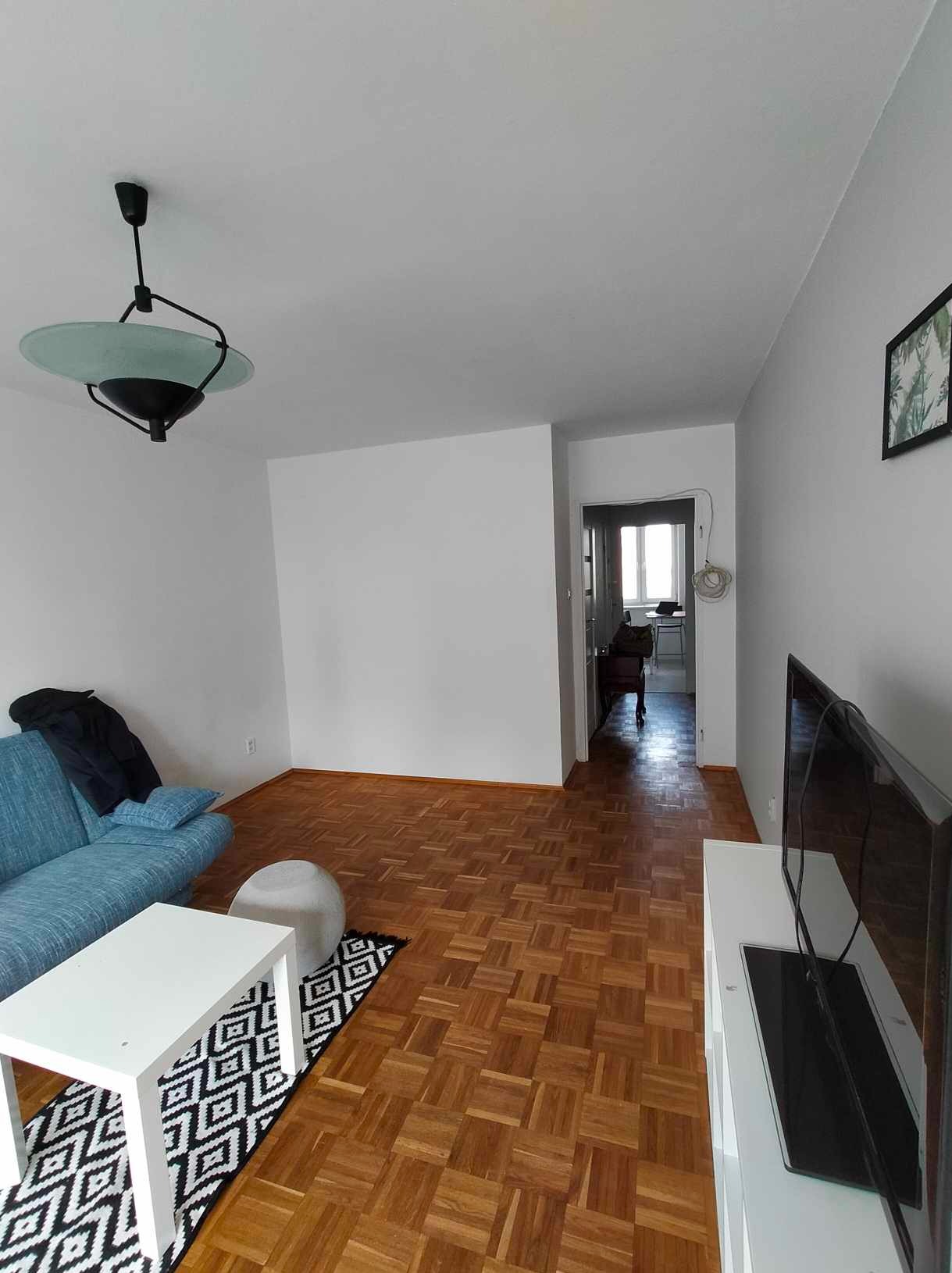 Aranżacja salonu w Warszawie: parkiet w jodełkę, niebieska sofa, biały stolik kawowy z geometrycznym dywanem. Widok na dalsze pomieszczenie.