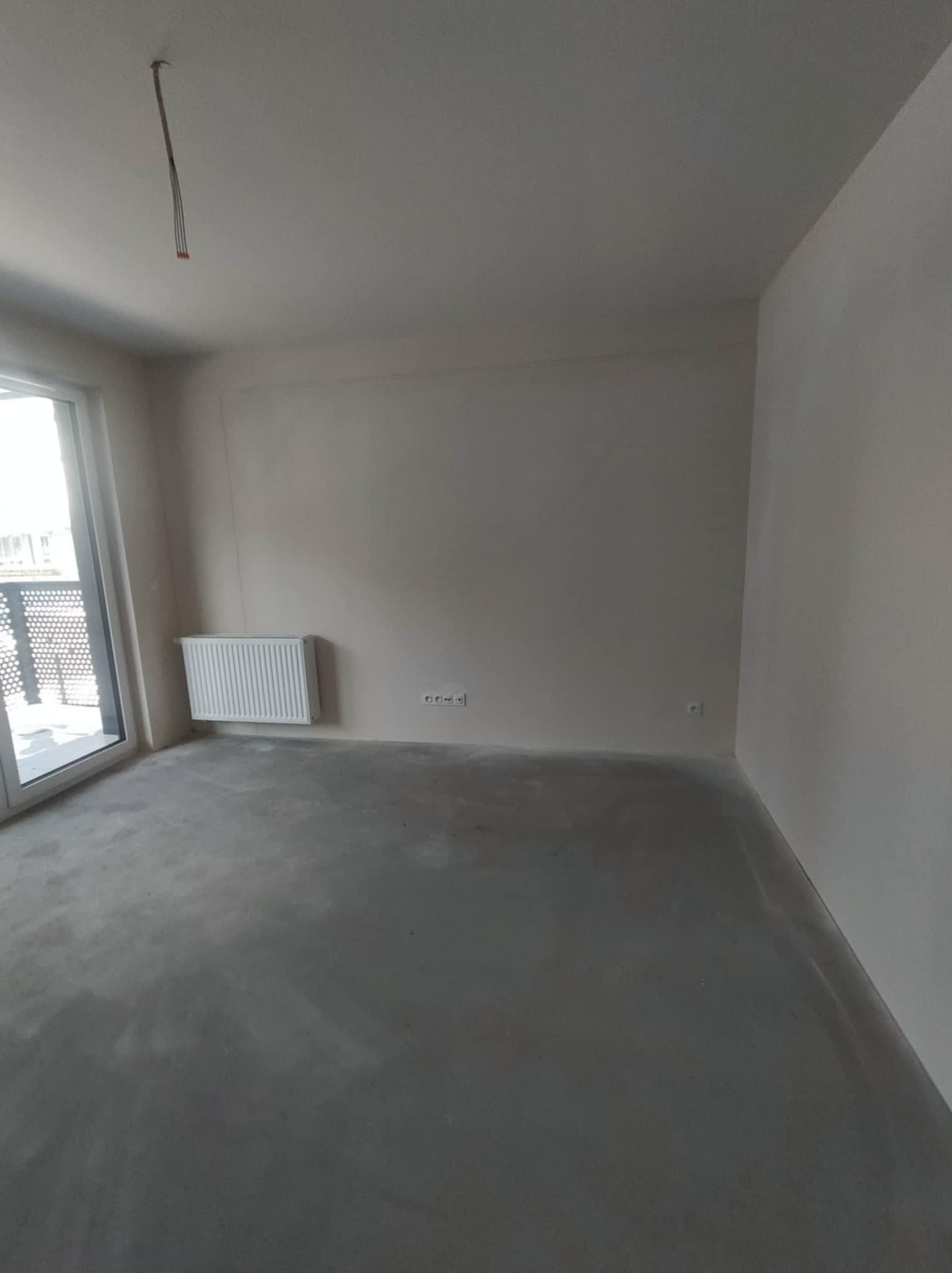 Surowy stan developerski mieszkania w Warszawie: betonowa podłoga, białe ściany, grzejnik i wystające kable. Widok z okna na balkon.