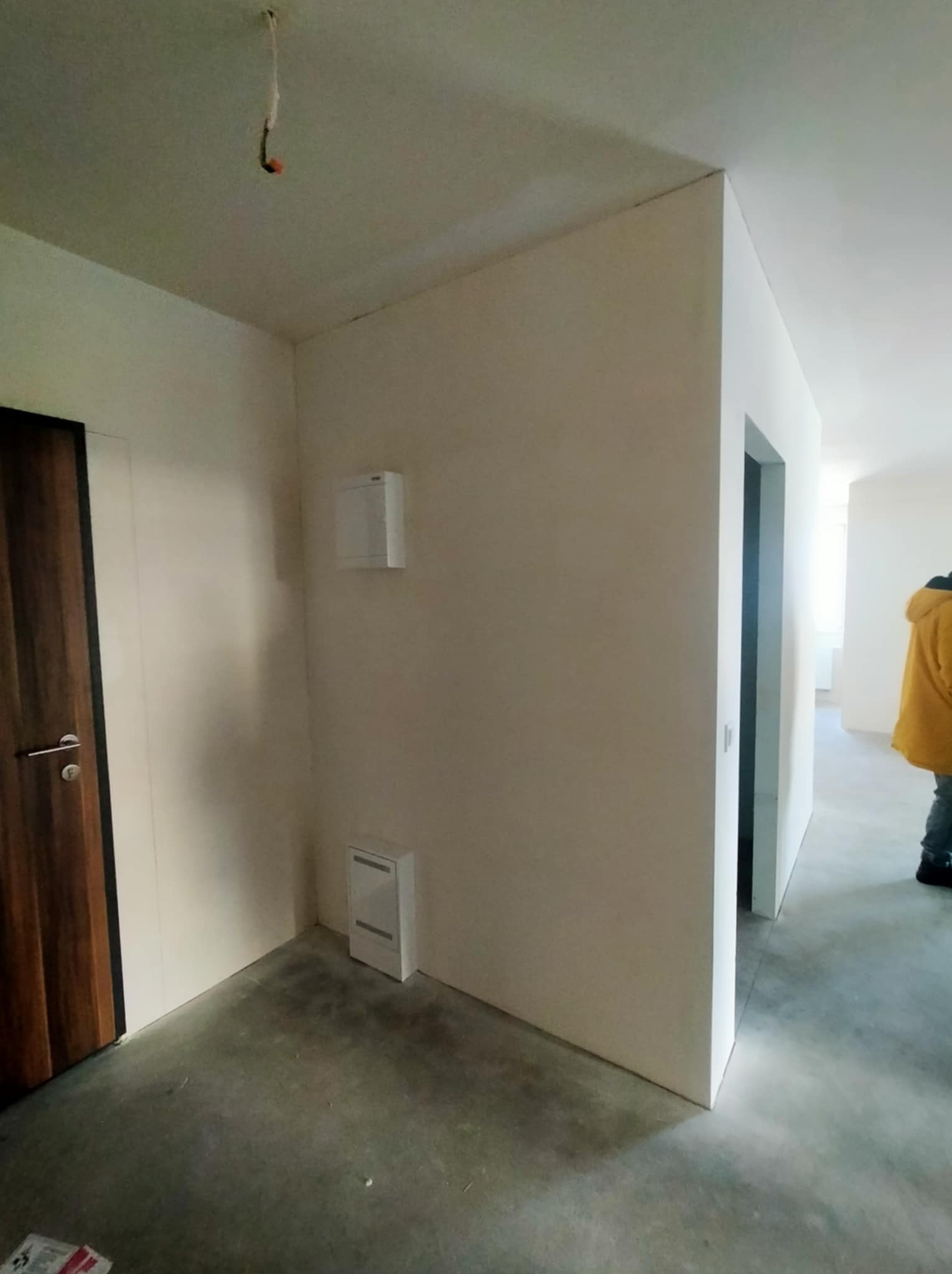 Wnętrze mieszkania w stanie deweloperskim: surowe ściany, betonowa podłoga, widoczne instalacje. Drzwi z ciemnego drewna i fragment osoby w żółtej kurtce w tle.