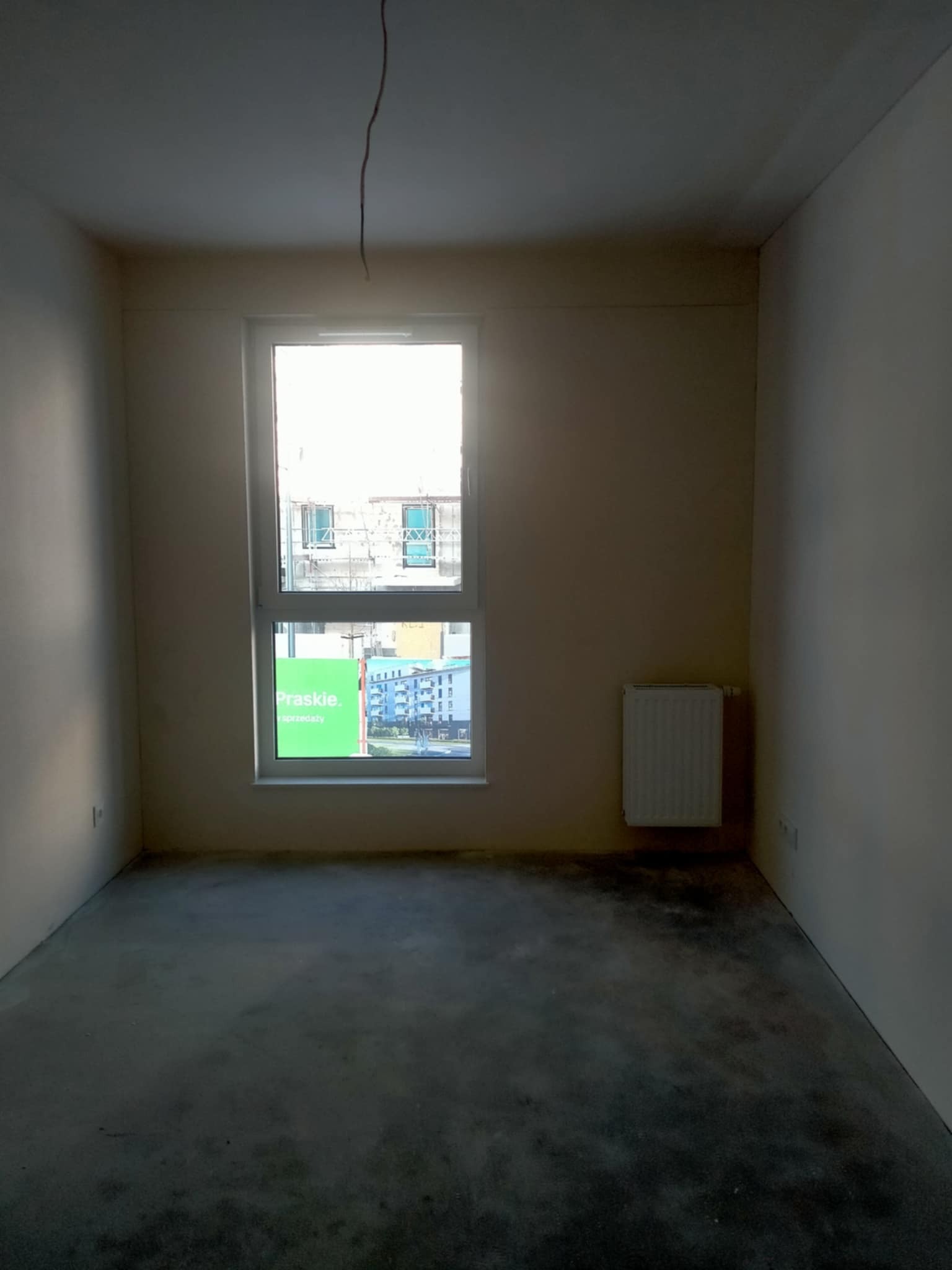 Surowe wnętrze pokoju z oknem i grzejnikiem. Widoczne ślady po pracach wykończeniowych na podłodze. Kabel zwisa z sufitu. Okno z widokiem na budowę.