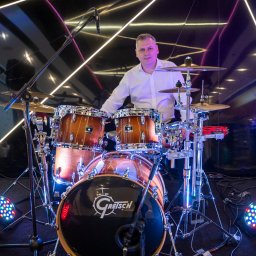 JakByCo - Perkusista w białej koszuli za zestawem perkusyjnym marki Gretsch na scenie z neonowym oświetleniem w tle.