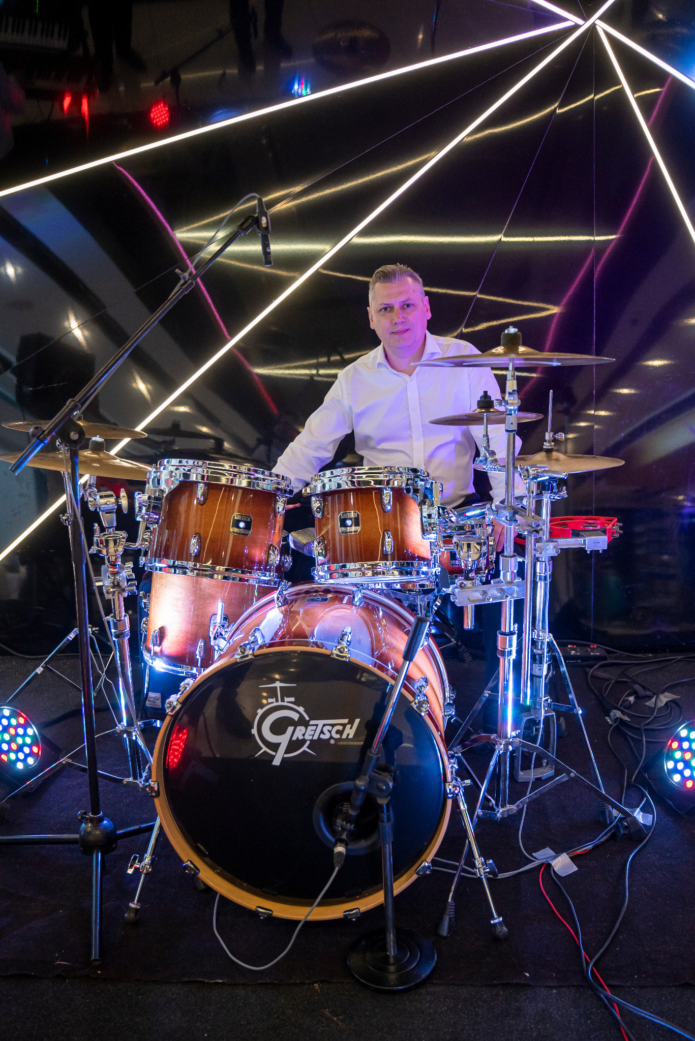 Perkusista w białej koszuli za zestawem perkusyjnym marki Gretsch na scenie z neonowym oświetleniem w tle.