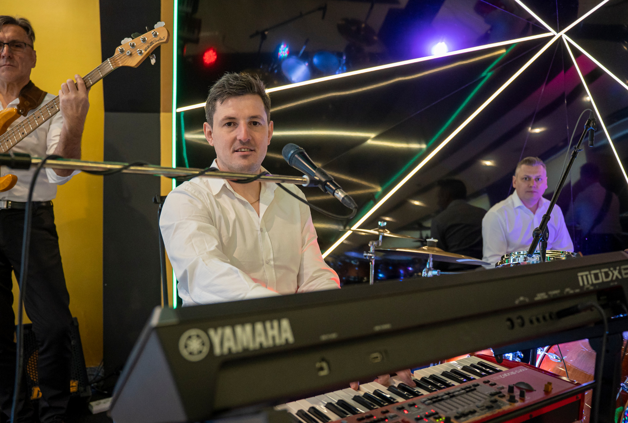 Muzycy w białych koszulach podczas występu: klawiszowiec z mikrofonem, basista z gitarą basową i perkusista w tle na scenie z oświetleniem.