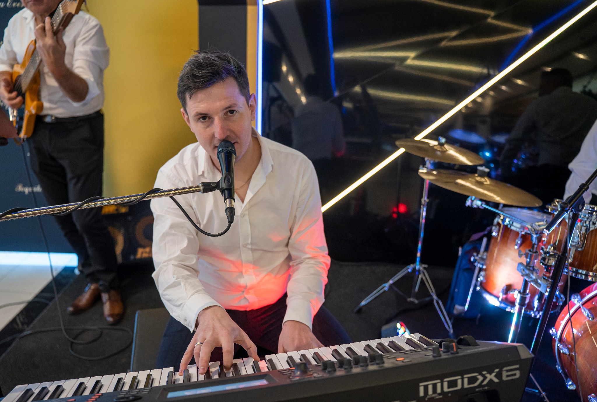 Wokalista grający na keyboardzie Yamaha MODX6, z perkusją i gitarzystą w tle, oświetlenie sceniczne w kolorach żółtym i niebieskim.