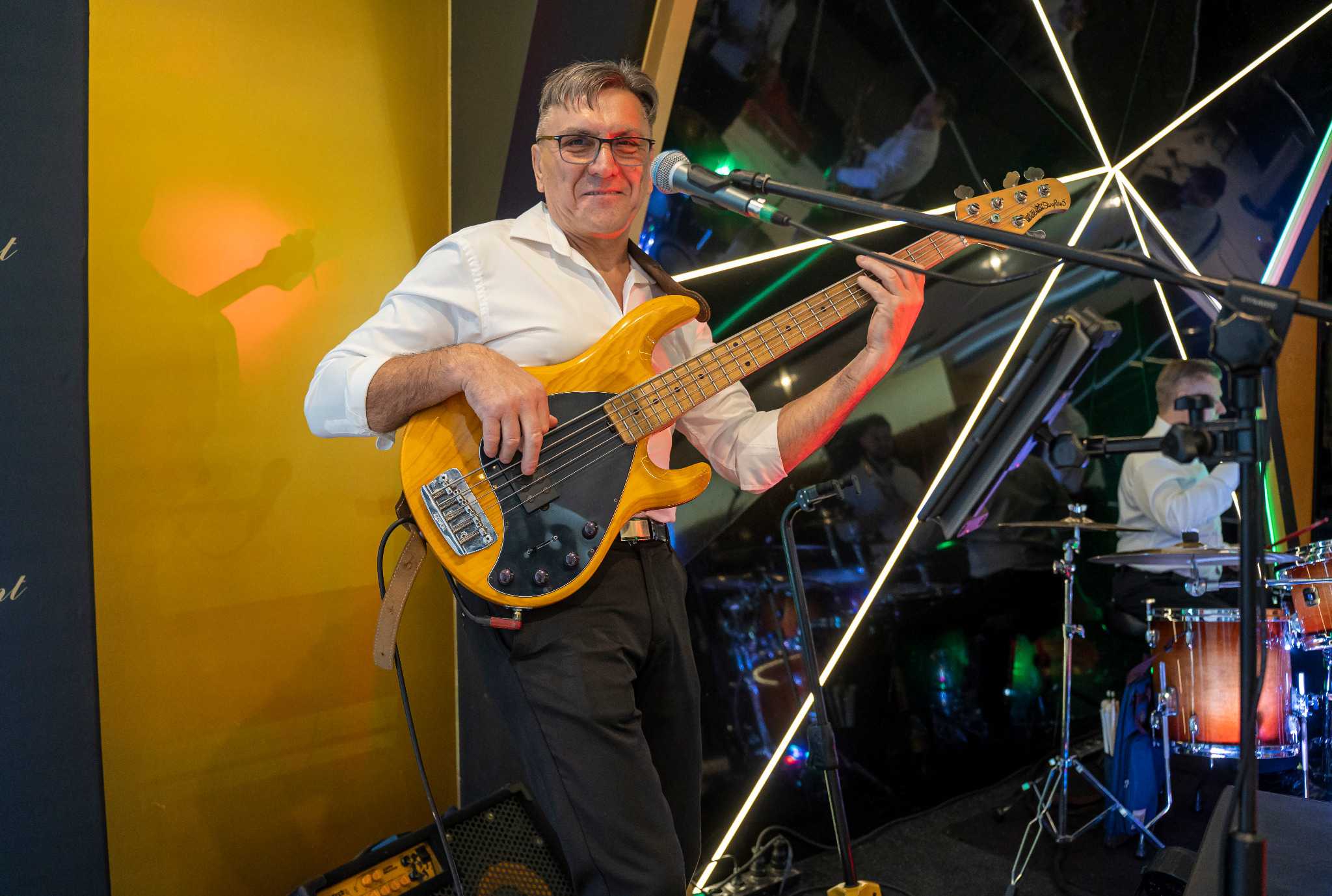 Uśmiechnięty basista w okularach gra na żółtej gitarze basowej podczas występu na scenie z perkusją i oświetleniem.