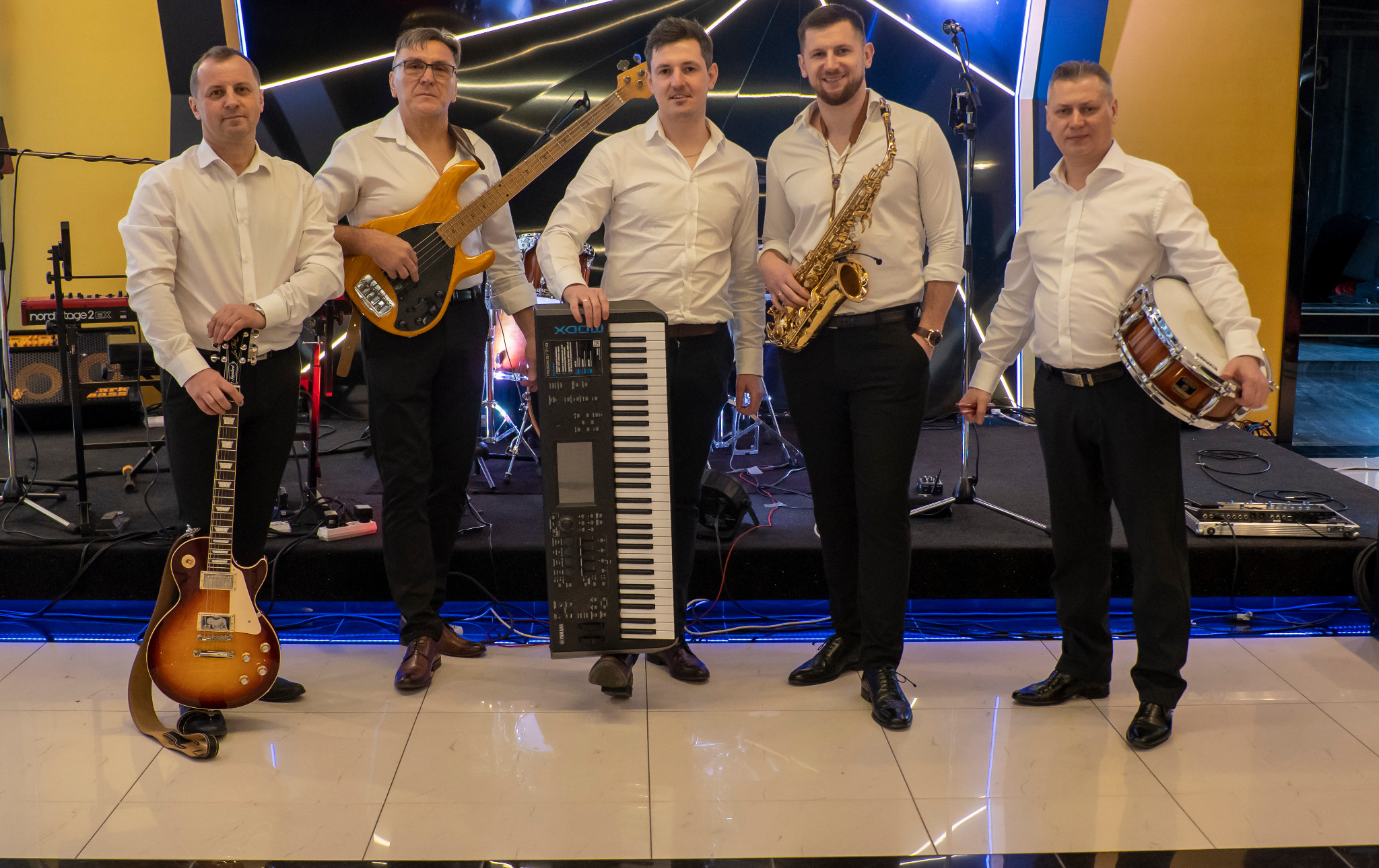 Pięcioosobowy zespół w białych koszulach prezentuje instrumenty: gitara elektryczna, gitara basowa, keyboard Yamaha MODX7, saksofon i bęben. Instrumenty trzymane w rękach, ustawione na scenie...