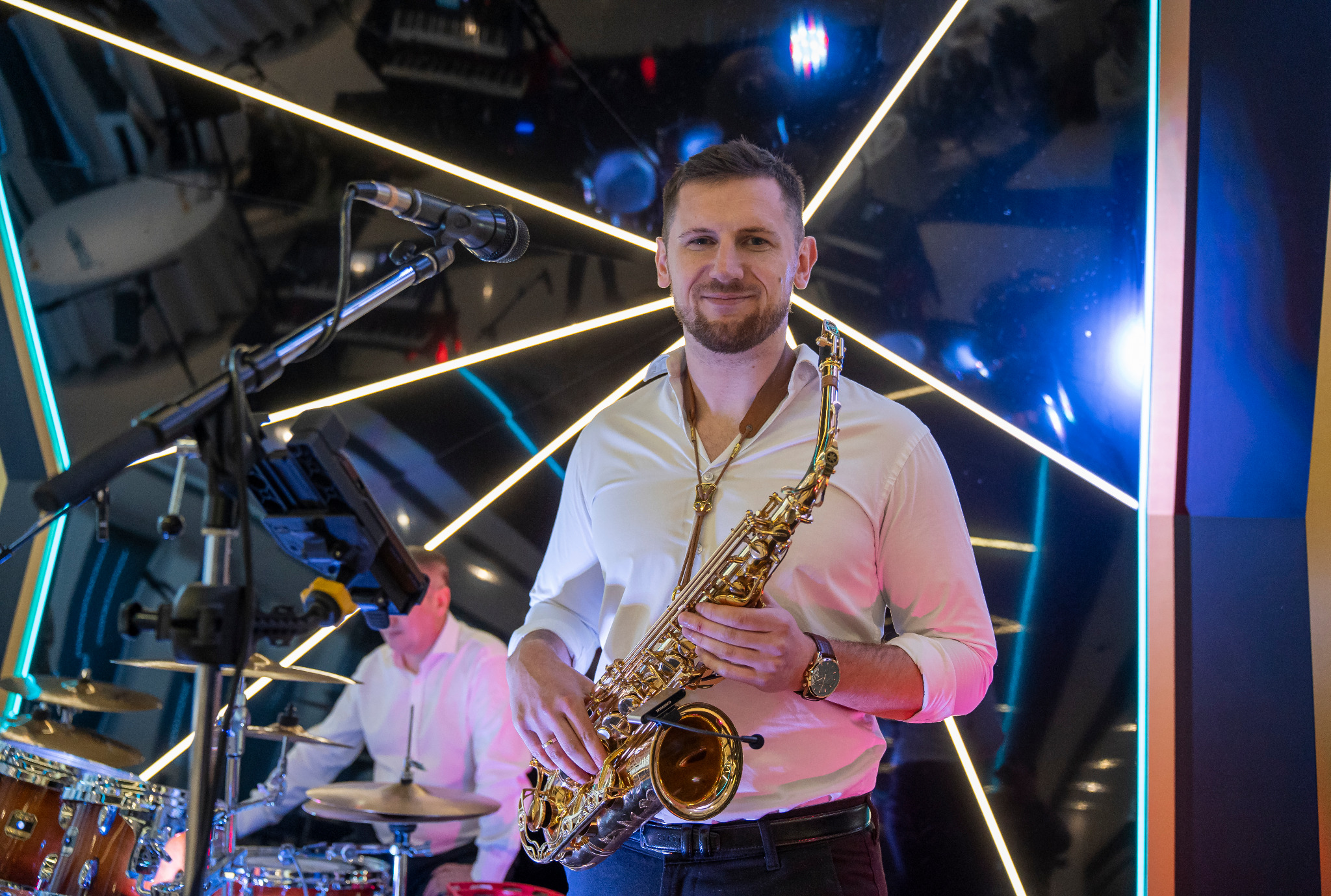 Uśmiechnięty saksofonista w białej koszuli, z saksofonem na tle perkusji i neonowego oświetlenia scenicznego.