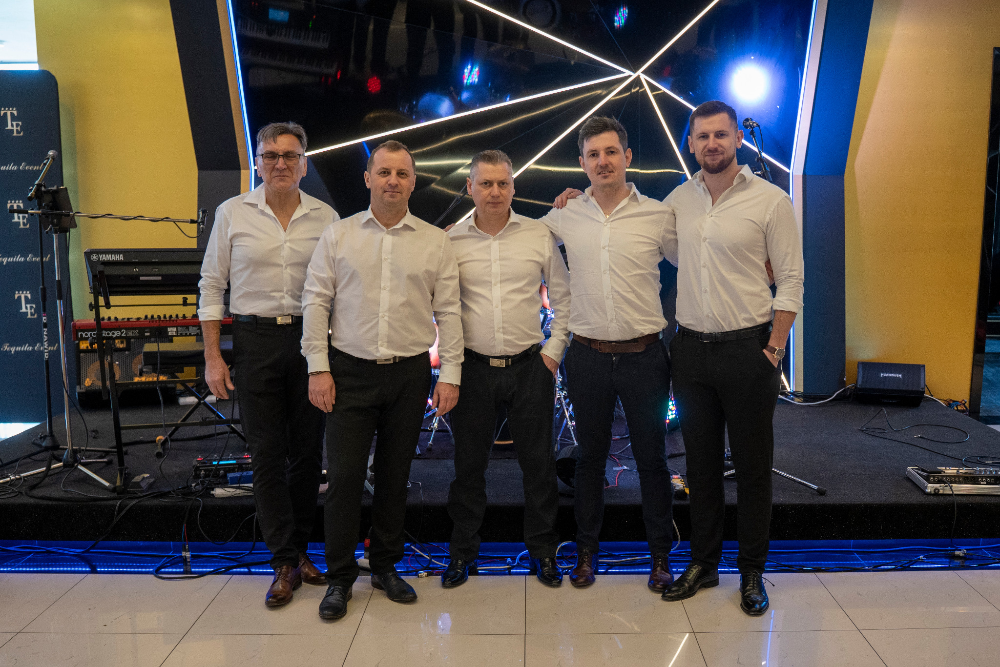 Pięciu mężczyzn w białych koszulach i czarnych spodniach pozuje na scenie przed instrumentami muzycznymi i dekoracjami świetlnymi, w tym keyboard Yamaha i oświetlenie LED.