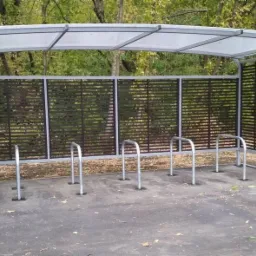 Zadaszona wiata rowerowa z metalową konstrukcją, ażurowymi ścianami i przezroczystym dachem, umieszczona na betonowym podłożu w otoczeniu drzew.