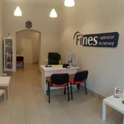 Jasne wnętrze biura operatora bankowego Fines z białymi meblami, krzesłami z czerwonymi oparciami, logo firmy na ścianie i zegarem nad wejściem.