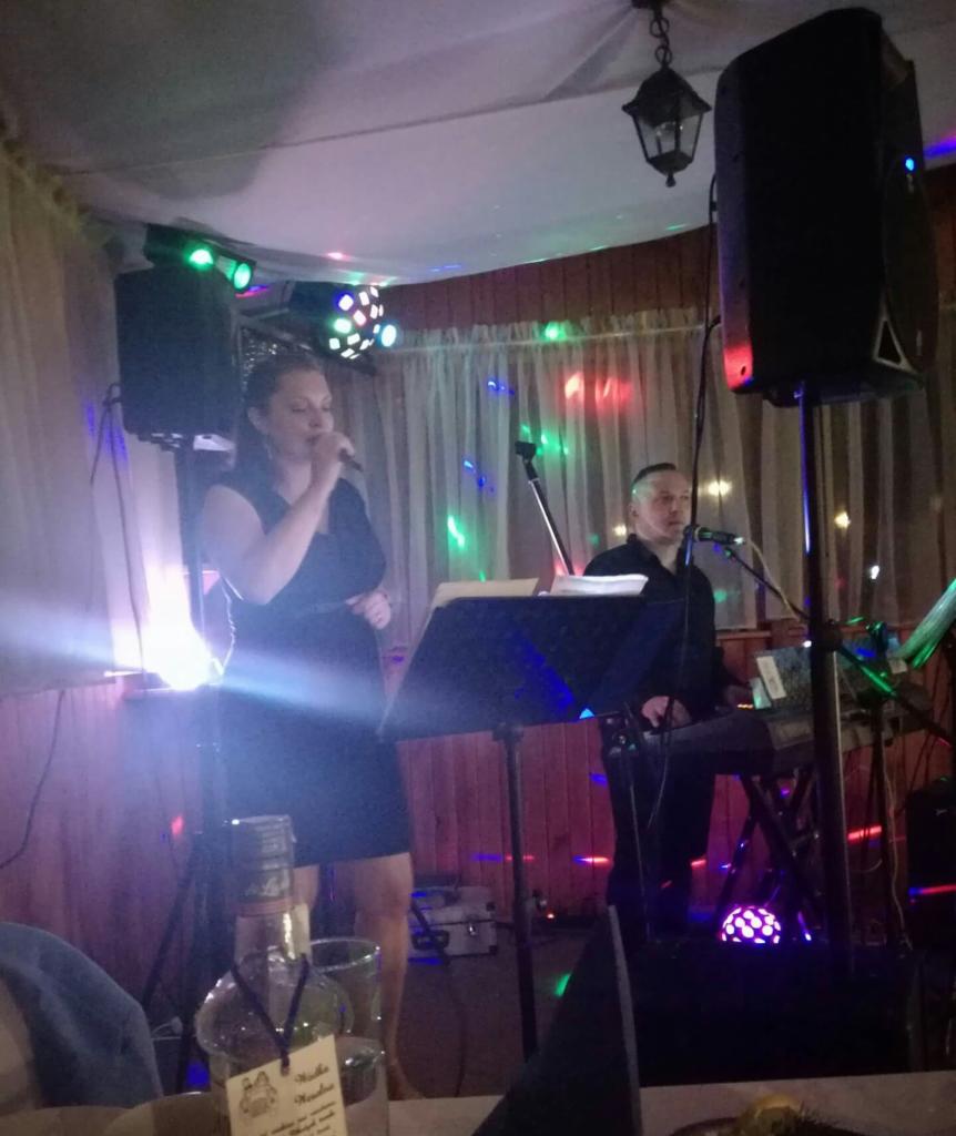 Wokalistka w czarnej sukience śpiewa do mikrofonu na tle dekoracji świetlnych, obok klawiszowiec gra na keyboardzie, widoczny stolik z butelką i kieliszkami w pierwszym planie.