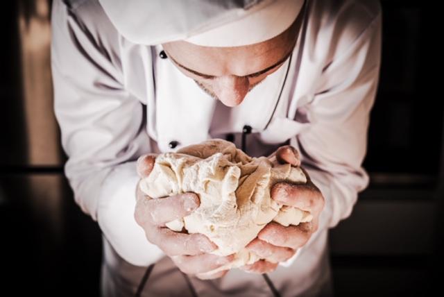 Szef kuchni w białym uniformie zagniata ciasto w dłoniach, pochylony nad nim, z widocznymi drobnymi zmarszczkami na czole, w tle błyszcząca powierzchnia sprzętu kuchennego.