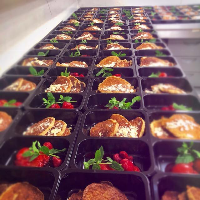 Rząd pojemników z gotowymi posiłkami: naleśniki z białym serem i migdałami, podane z sosem malinowym i świeżą miętą.