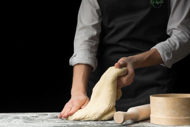 Kucharz w czarnym fartuchu ugniata ciasto na oprószonej mąką powierzchni, obok drewniany wałek i sito, ciemne tło.