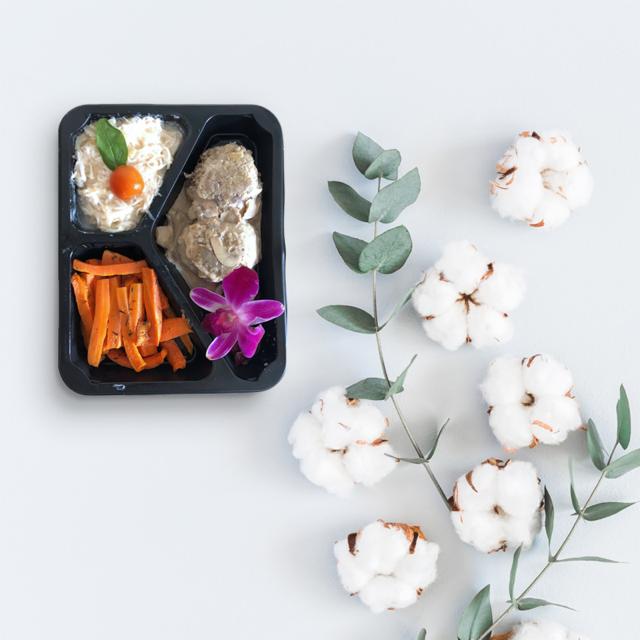 Czarna tacka z trzema przegródkami zawierająca porcje: makaronu z sosem i pomidorkiem cherry, klopsików w sosie grzybowym z plasterkiem migdała oraz marchewki glazurowanej, udekorowana fioletowym...