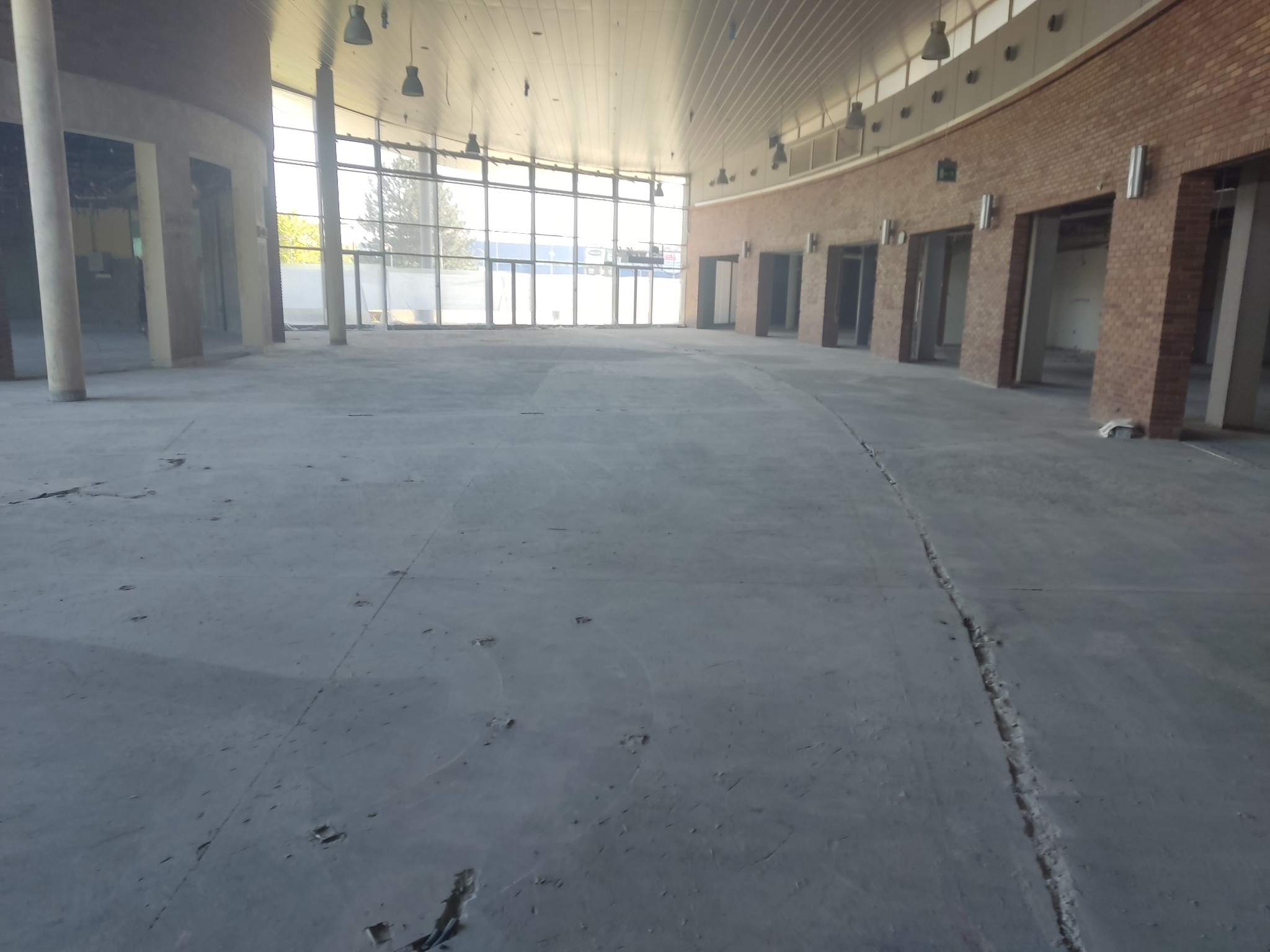 Przestronne wnętrze hali po rozbiórce, widoczne ślady po ścianach i instalacjach, duże okna z widokiem na zewnątrz, surowy beton na podłodze z pęknięciami.